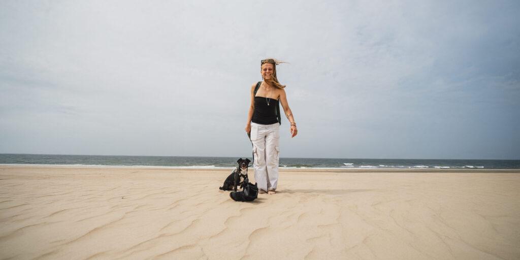 “Vrouw staat op een leeg zandstrand met een zwarte hond, uitkijkend over de zee onder een bewolkte hemel.”