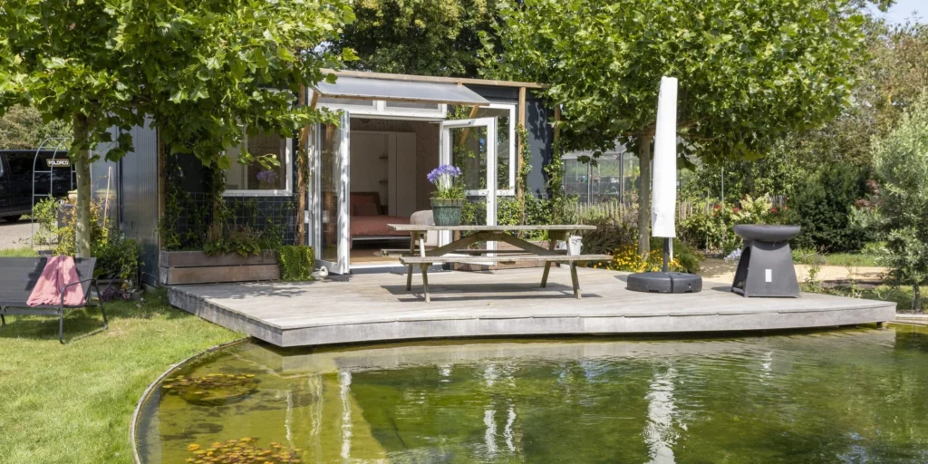 Een kleine houten tuinunit met openslaande deuren staat op een houten vlonder aan een vijver, omringd door groen, een picknicktafel en een parasol.