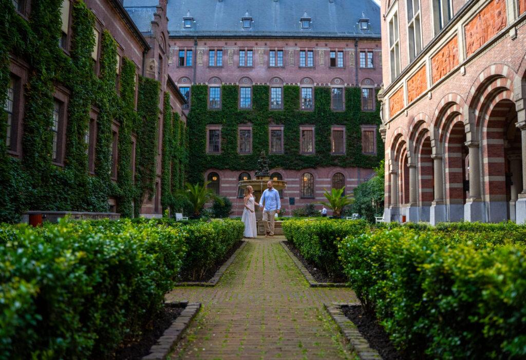 Bruidspaar loopt samen door een symmetrische tuin bij een historisch gebouw tijdens hun bruiloft.