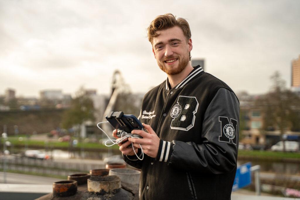 Een jonge man met een zwarte varsity-jas staat bij een reling aan het water en houdt een camera met kabels in zijn handen terwijl hij naar de camera kijkt.
