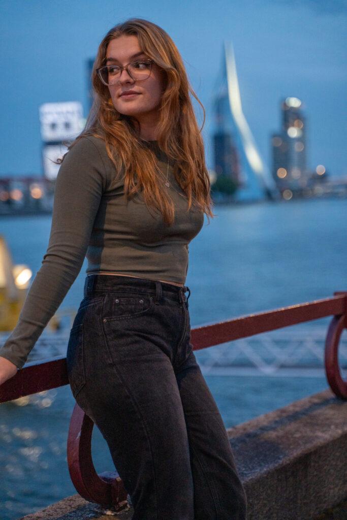 Vrouw poseert ontspannen met de skyline van Rotterdam op de achtergrond tijdens het blauwe uur.