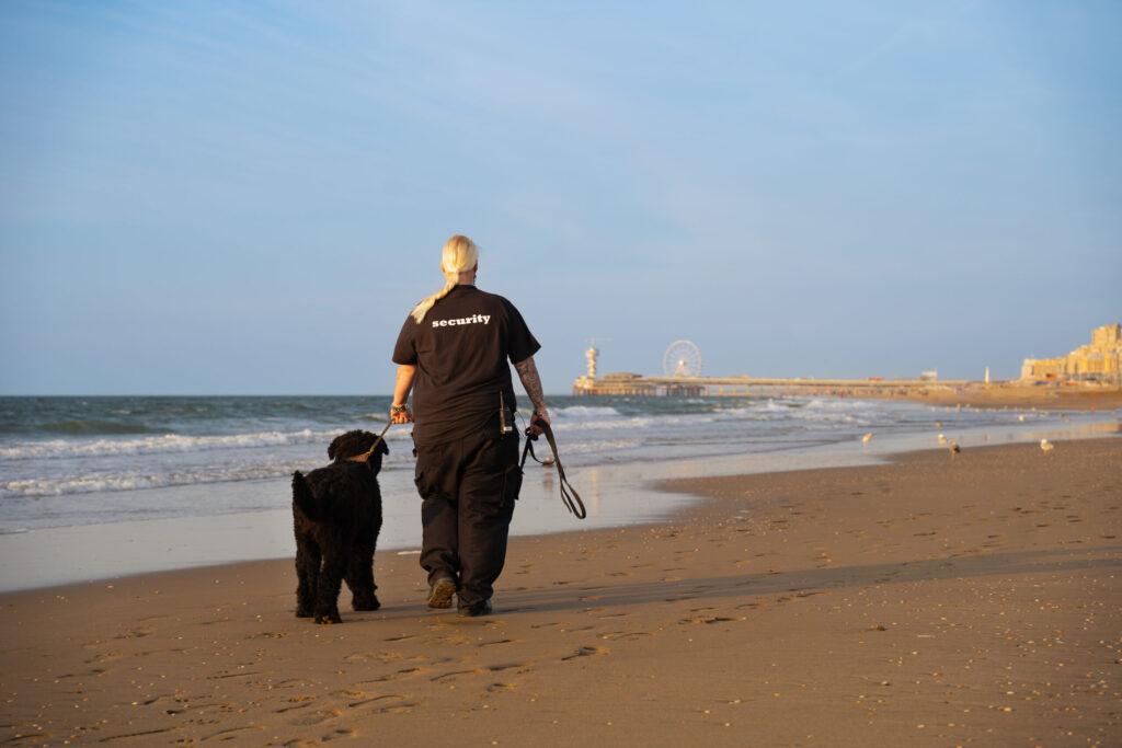 Een vrouwelijke beveiligingsmedewerker in zwart uniform loopt met een grote zwarte hond langs de waterlijn op het strand, met een pier en een reuzenrad op de achtergrond.