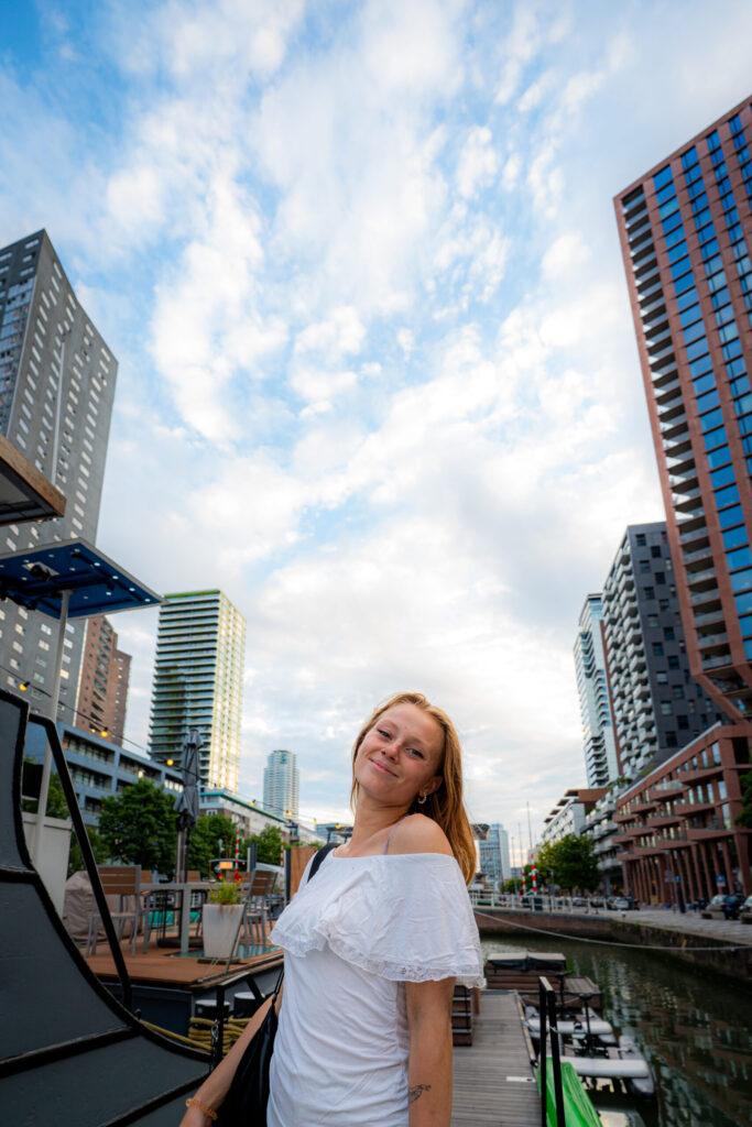 Vrouw in een witte off-shoulder jurk staat glimlachend aan de Wijnhaven in Rotterdam, met moderne hoogbouw en boten op de achtergrond.