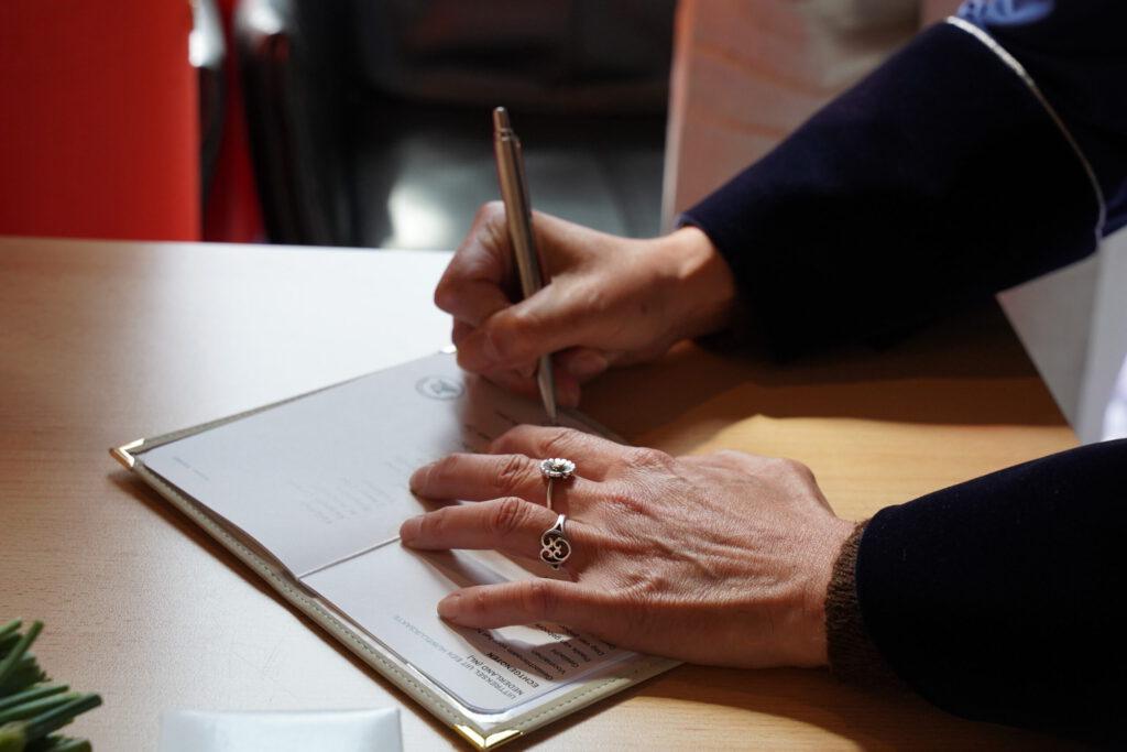 Close-up van een hand met een ring die een officiële huwelijksakte ondertekent met een pen op een houten tafel.