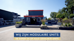 Een zwarte modulaire Foldaco-unit met rode dakrand en glazen entree, geplaatst op een bedrijventerrein met sierbestrating en groen eromheen.