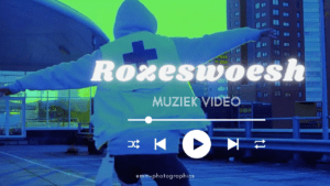 Een persoon met uitgestrekte armen staat op een stadsbrug bij nacht, met een fel groen-blauw kleurfilter en de tekst “Roxeswòesh – Muziek Video” als overlay.