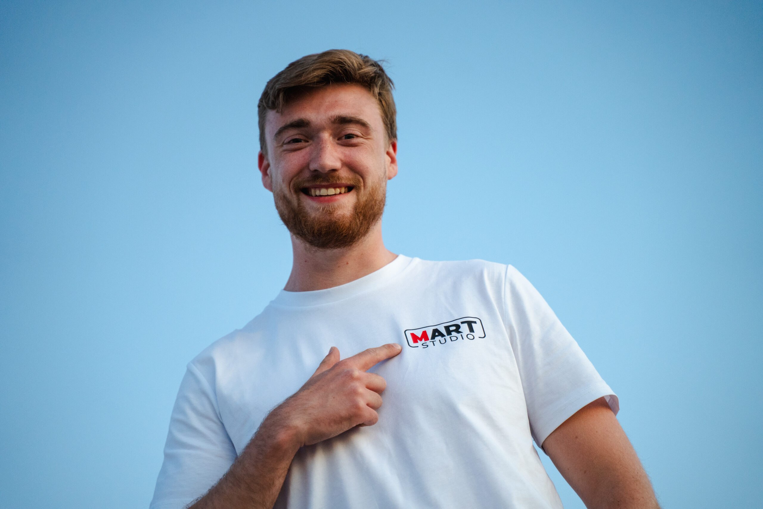 Man wijst naar Mart Studio logo op wit T-shirt tegen blauwe achtergrond