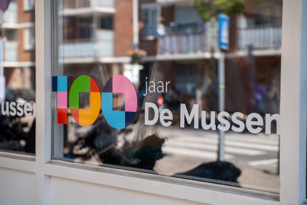100 jaar jubileumlogo van De Mussen op een raam van het buurtcentrum.