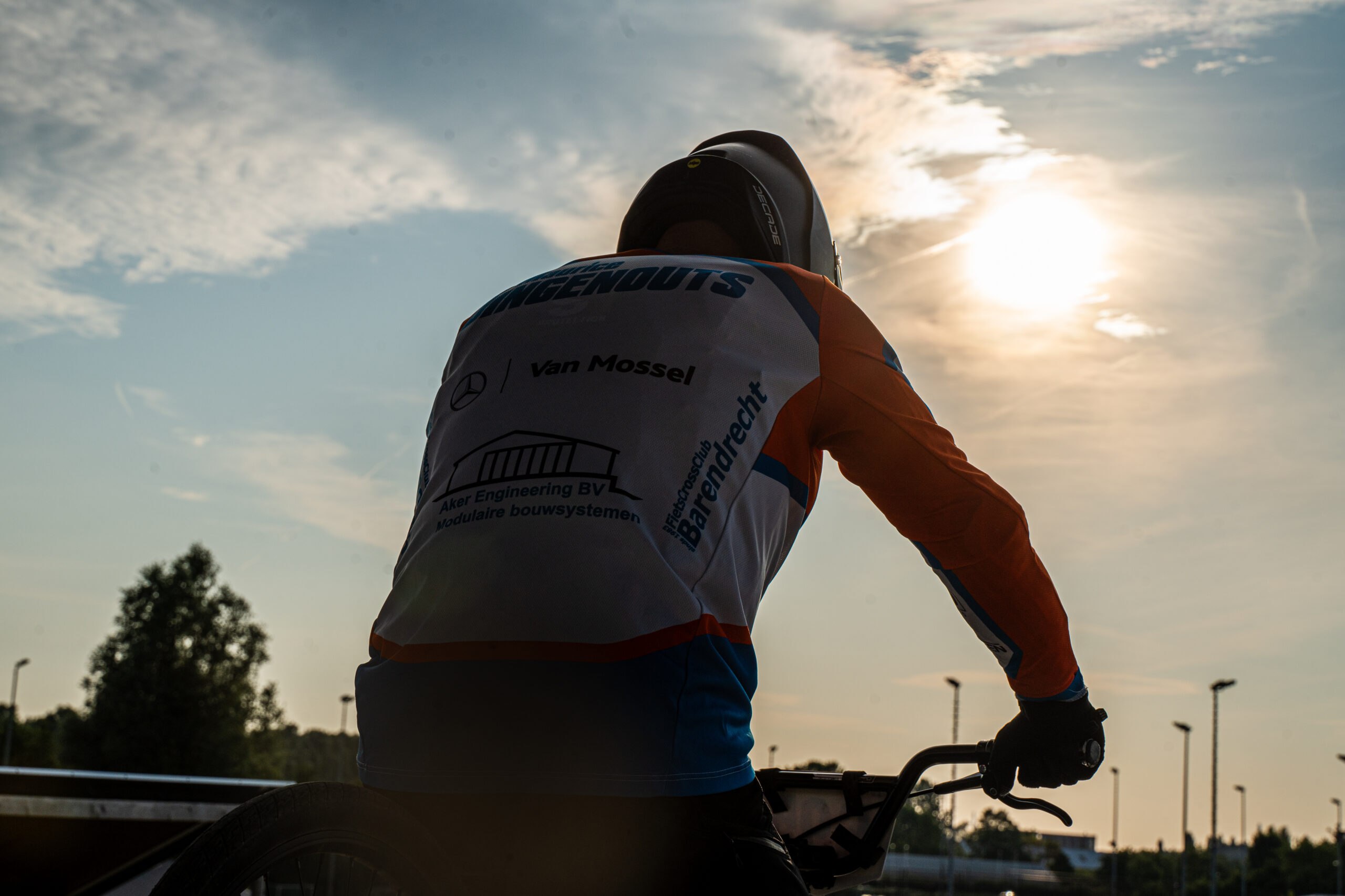 BMX-rijder kijkt richting de baan met de laagstaande zon op de achtergrond.