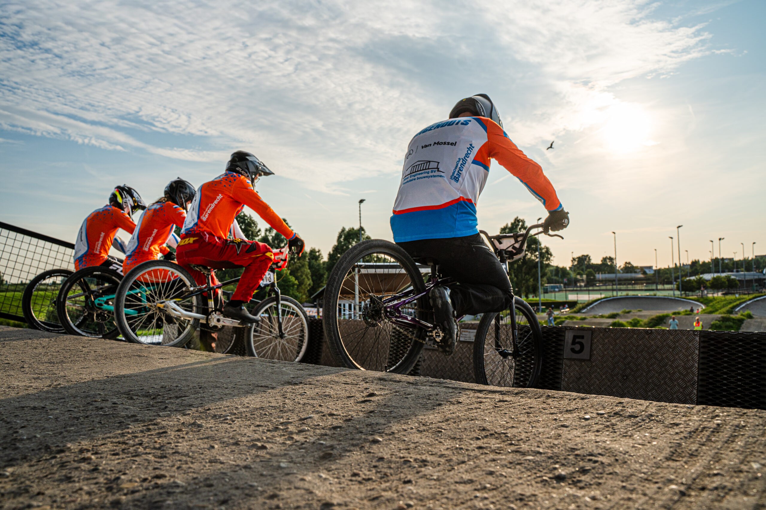 BMX-rijders wachten aan het starthek terwijl de zon laag aan de hemel staat.